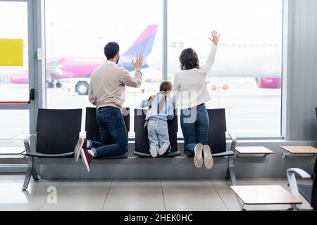 vista posteriore della famiglia che sventolano le mani mentre si guarda l'aeroplano attraverso la finestra in aeroporto Foto Stock