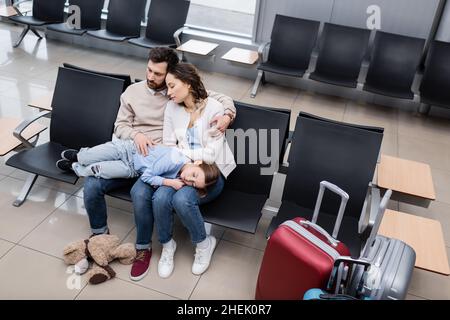 vista ad alto angolo della ragazza che dorme sulle ginocchia dei genitori stanchi nella lounge dell'aeroporto Foto Stock