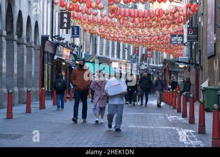 Londra, UK, 9 gennaio 2022: Su Lisle Street a Chinatown le persone indossano maschere per il viso anche all'aperto, in quanto i tassi omicron rimangono molto alti nella capitale nonostante i numeri si siano livellati negli ultimi giorni. Oltre ai turisti che passano accanto, un operatore di ristoranti in pantaloni check porta una scatola sotto le lanterne rosse cinesi. Anna Watson/Alamy Foto Stock