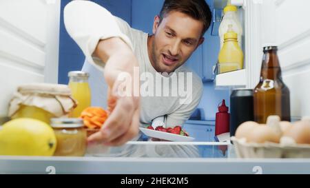 uomo gioioso che prende deliziosi cupcake e fragole dal frigorifero con bevande, uova e contenitori con cibo Foto Stock