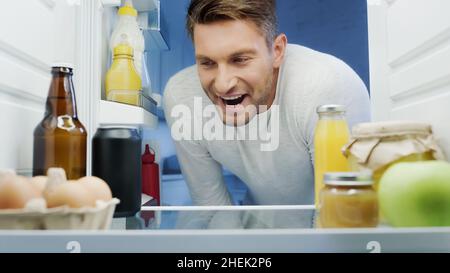 uomo emozionato che guarda in frigorifero con bevande, uova, vasetti e bottiglie con salse Foto Stock