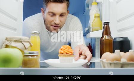 uomo contento che prende piatto con deliziosi cupcake dal frigorifero con bevande, uova, vasetti e bottiglie con salse Foto Stock