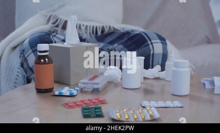 vista ritagliata dell'uomo malato seduto sul divano sotto coperta calda vicino al tavolo con medicamenti e tovaglioli di carta Foto Stock