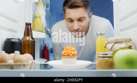 uomo che guarda deliziosi cupcake vicino a bevande, uova e salse in frigorifero Foto Stock