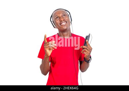 un bell'uomo in una t-shirt rossa in piedi su uno sfondo bianco che ascolta la musica con le cuffie utilizzando il cellulare e guardando verso l'alto sorridente. Foto Stock
