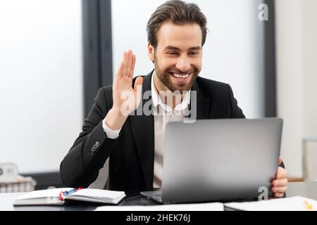 Sorridendo il giovane uomo d'affari con abbigliamento formale seduto alla scrivania in ufficio, studiando o lavorando online, avendo incontri virtuali, agitando allo schermo del laptop, il dipendente maschile partecipa a una conferenza online Foto Stock