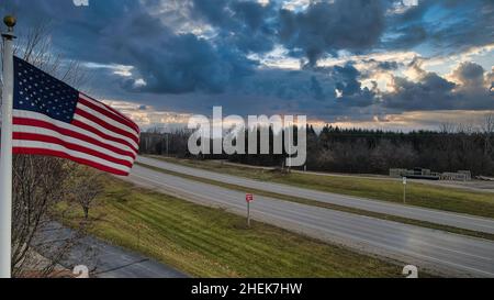 La bandiera americana si muove nel vento con le nuvole scure sopra. Una strada rurale vuota sullo sfondo con alberi. Foto Stock