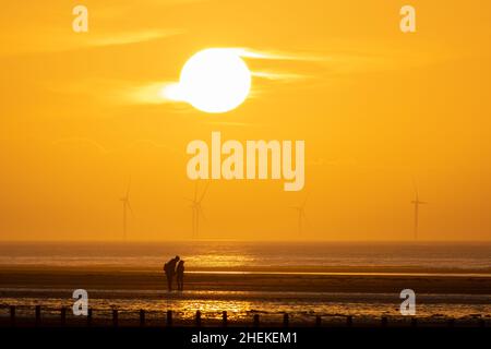 Ainsdale Beach, Southport, Merseyside, Regno Unito. 11th Jan 2022. Il sole si prepara a tramontare dietro turbine eoliche lontane mentre il cielo si allontano, con l'alta pressione che si muove e le condizioni nel Nord Ovest che saranno sistemate per tutta la settimana. Una coppia si erge sotto il sole. Credit: Callum Fraser/Alamy Live News Foto Stock