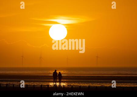 Ainsdale Beach, Southport, Merseyside, Regno Unito. 11th Jan 2022. Il sole si prepara a tramontare dietro turbine eoliche lontane mentre il cielo si allontano, con l'alta pressione che si muove e le condizioni nel Nord Ovest che saranno sistemate per tutta la settimana. Una coppia si erge sotto il sole. Credit: Callum Fraser/Alamy Live News Foto Stock