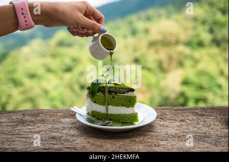 Tenere una tazza versando salsa di tè verde su torta di tè verde su un bar in legno nella piantagione Foto Stock