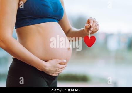 Giovane donna incinta in abbigliamento sportivo che tiene il cuore rosso appeso sul bastone in mano e carezzando il ventre. Foto Stock