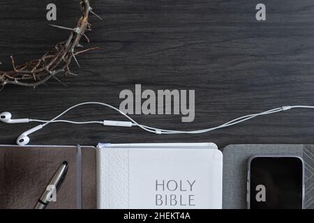 Bordo della bibbia bianca e diari grigi con corona di spine e telefono cellulare e auricolari su sfondo di legno scuro con spazio copia Foto Stock