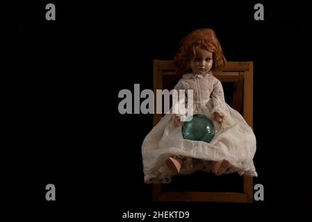 Bambola in porcellana vintage con capelli rossi e occhi verdi che indossa un abito bianco, siede su una sedia di legno, su sfondo nero con una palla di vetro in grembo. Foto Stock