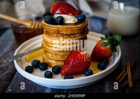 Vista chiara della pila di frittelle con bastone di miele, fragola, bacca blu e panna frusta in cima nel piatto, delizioso dessert con latte per la colazione. Foto Stock