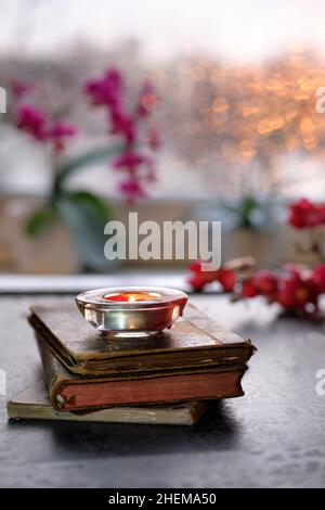 Inverno con candela su pila di vecchi libri Sunset window con arancio bagliore, rosa e fucsia orchidea e fiori magnolia. Candele aromatiche, luce del tè Foto Stock