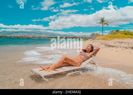 Donna vacanza spiaggia relax in bikini sdraiati su lettino sole prendere il sole con sedia nelle onde dell'oceano in Hawaii. USA Isola Hawaiiana Foto Stock