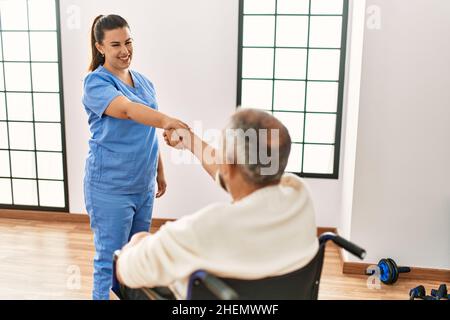 Uomo anziano seduto su sedia a rotelle e infermiera geriatrica che fa accordo di scuotimento della mano, ringraziando per l'aiuto Foto Stock