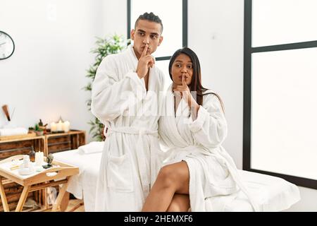 Giovane coppia latina con asciugamano in piedi al centro di bellezza chiedendo di essere tranquillo con le dita sulle labbra. Silenzio e concetto segreto. Foto Stock