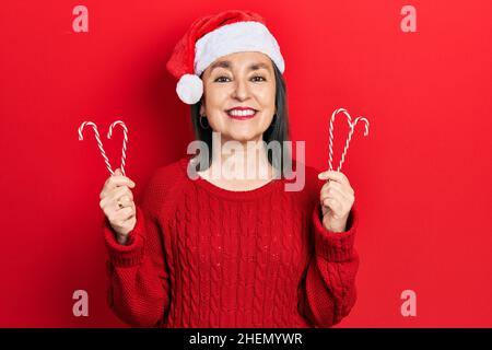 Donna ispanica di mezza età che indossa il cappello di natale che tiene caramelle sorridendo con un sorriso felice e fresco sul viso. Mostrando i denti. Foto Stock