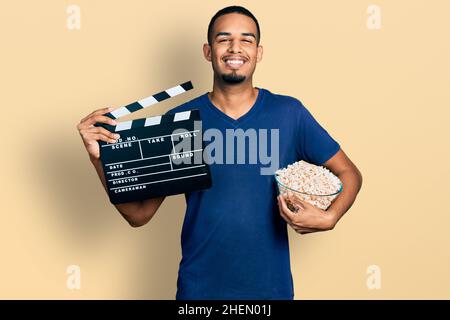 Giovane uomo afroamericano che mangia popcorn regge film clapboard sorridendo con un sorriso felice e fresco sul viso. Mostrando denti. Foto Stock