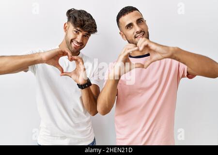 Giovane coppia gay in piedi su sfondo isolato sorridendo nell'amore facendo la forma del simbolo del cuore con le mani. Concetto romantico. Foto Stock