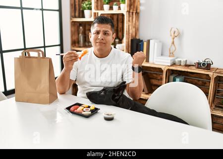 Giovane ispanico che mangia sushi usando bacchette urlando orgoglioso, celebrando la vittoria e il successo molto eccitato con le braccia sollevate Foto Stock