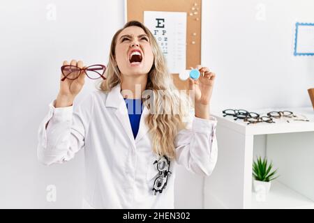 Giovane bella donna optician che tiene gli occhiali e le lenti a contatto arrabbiato e pazzo urlando frustrati e furiosi, urlando con rabbia guardando in su. Foto Stock