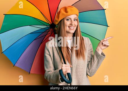 Giovane irlandese in stile francese che tiene un ombrello colorato sorridendo felice di puntare con mano e dito a lato Foto Stock