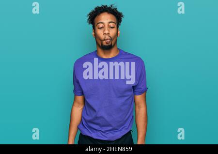 Giovane afroamericano con barba con maglietta viola casual che fa il viso del pesce con le labbra, gesto pazzo e comico. Espressione divertente. Foto Stock