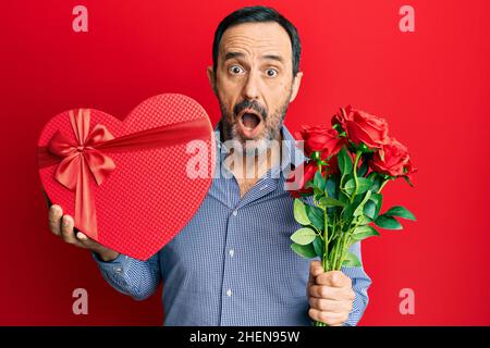 Uomo ispanico di mezza età in possesso di San Valentino regalo e fiori impauriti e sconvolti con sorpresa e stupito espressione, paura e volto eccitato. Foto Stock