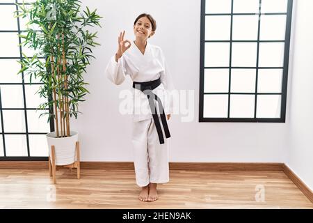 Giovane ispanica ragazza che indossa kimono karate e cintura nera sorridendo positivo facendo segno ok con mano e dita. Espressione di successo. Foto Stock