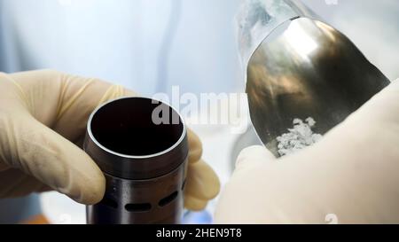 Primo piano delle mani in guanti medici che mettono polvere chimica dal cucchiaio per sostanze chimiche al piccolo contenitore di metallo in laboratorio. Mani inserimento del reagente bianco Foto Stock