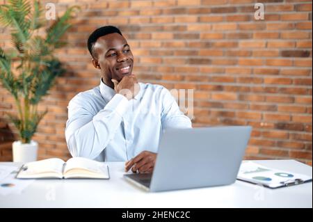 Orgoglioso uomo d'affari afro-americano di successo, top manager, capo reparto, si siede sul posto di lavoro in un ufficio moderno, guarda con attenzione al lato, sorride, medita un progetto, sogni di una vacanza Foto Stock