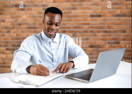 Un uomo d'affari afro-americano focalizzato che scrive note nel notebook durante il webinar online, sta migliorando le qualifiche. Smart black maschio studente studiare online, ascoltare lezioni online, utilizzare il laptop Foto Stock
