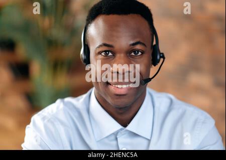 Primo piano di un piacevole uomo afroamericano, con cuffie, esperto, consulente o operatore di hotline, guarda la fotocamera, sorride amichevole. Concetto di servizio di supporto, helpline, comunicazione a distanza Foto Stock