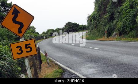 Un angolo stretto e tortuosa strada che arriva su una costa occidentale NZ strada. il segno 35kph è la velocità consigliata per affrontare le curve in modo sicuro Foto Stock