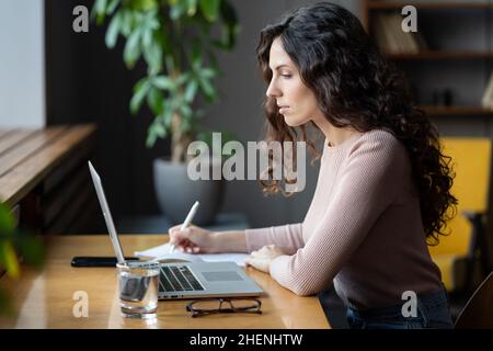Giovane donna d'affari seriamente concentrata che guarda il webinar aziendale sul notebook e prende appunti sul notebook Foto Stock