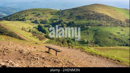 Una panca di legno e punto di vista dalla cima più alta di Malvern, guardando verso Table Hill e North Hill coperto di verde lussureggiante erba, all'inizio di giugno soleggiato su Foto Stock