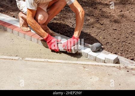 Un giovane lavoratore stende le lastre di pavimentazione in una luminosa giornata estiva di sole sulla base sabbiosa preparata di un percorso del parco in costruzione. Spazio di copia. Foto Stock