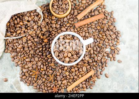 molti chicchi di caffè sono sparsi su un tavolo di marmo, in una tazza di caffè e in una borsa. bastoncini di cannella e stelle di anice si trovano sui grani. vista dall'alto Foto Stock