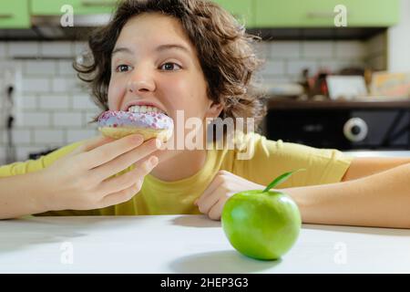 Bambino adolescente triste di fronte ad una scelta fra la ciambella e la mela verde. Ragazza teenage con il viso triste, livises dalla malnutrizione sotto i suoi occhi, è sulla dieta Foto Stock