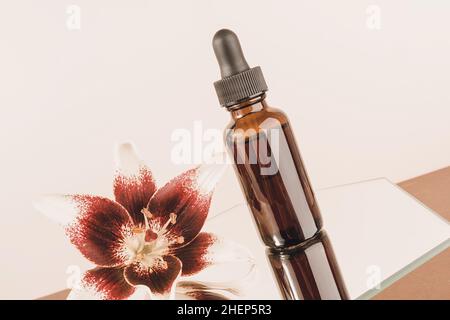 Una bottiglia di contagocce in vetro e bellissimo fiore sullo specchio, sfondo marrone. Natural Organic Spa concetto cosmetico. Vista frontale. Foto Stock