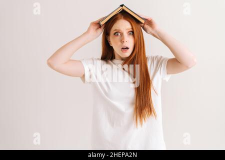 Ritratto dello studio di giovane studentessa stressata che copre la testa con libro che guarda via, in piedi su sfondo bianco isolato. Foto Stock