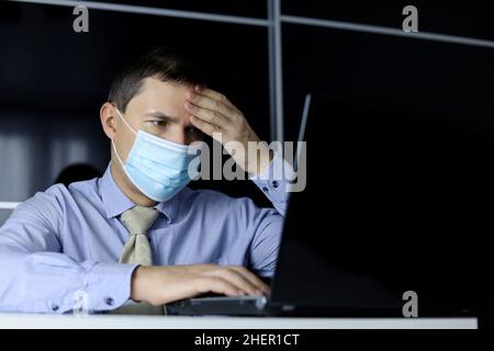 Coronavirus e sintomi di febbre, uomo in maschera facciale e vestiti da ufficio mettere la sua mano sulla fronte seduta al computer portatile. Lavoratore di ufficio malato Foto Stock