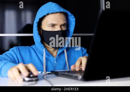 Uomo in maschera e felpa con cappuccio blu seduto al computer portatile. Concetto di lavoro di ufficio di sicurezza durante il coronavirus, il cyber crimine, l'hacking e la tecnologia Foto Stock