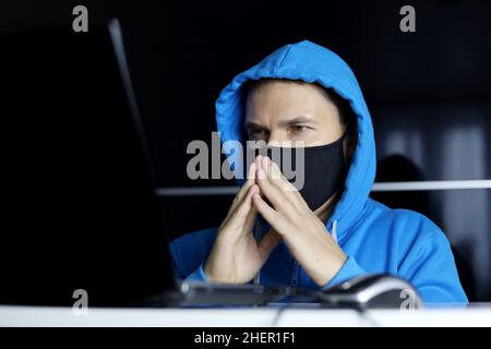 Uomo in maschera e felpa con cappuccio blu seduto al computer portatile con un'espressione del volto premurosa. Concetto di lavoro di sicurezza durante il coronavirus, cyber-crimine, hacking Foto Stock