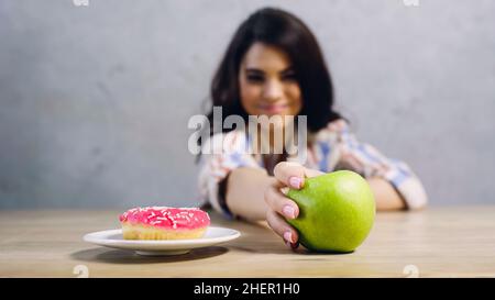 donna sfocata che prende mela verde vicino ciambella su grigio,immagine di scorta Foto Stock