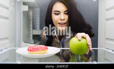 donna felice che prende mela verde vicino ciambella in frigorifero, immagine di scorta Foto Stock