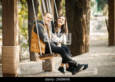 Ritratto di giovane coppia felice in sella ad un altalena in autunno in abiti caldi in colori base in un parco all'aperto. Foto di alta qualità Foto Stock