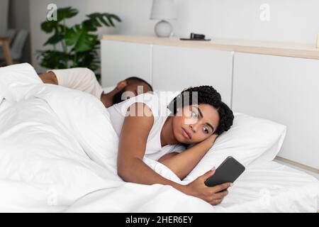 Triste giovane afroamericana donna guarda smartphone sdraiati su comodo letto nascondere imbrogliare Foto Stock